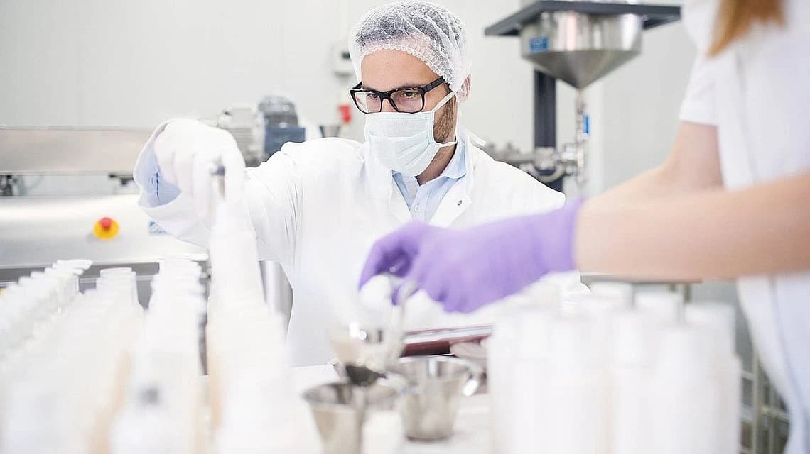 Two scientists in lab coats and gloves examine a bottle in a laboratory setting.