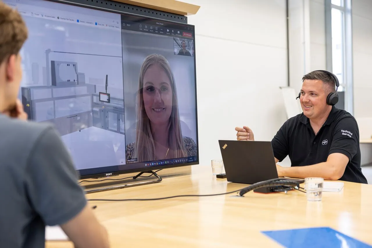 Two people at a table with a laptop and a TV, engaged in a discussion about the Körber Academy platform.