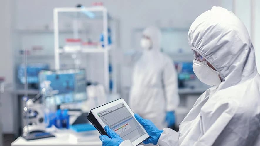 A person wearing a lab coat and gloves is holding a tablet, illustrating the use of CDMOS software in digital pharma.