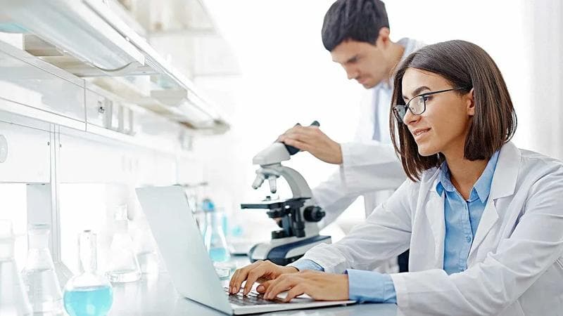 Woman in glasses, looking at a computer and wearing a lab coat. In the background, a man using a microscope.