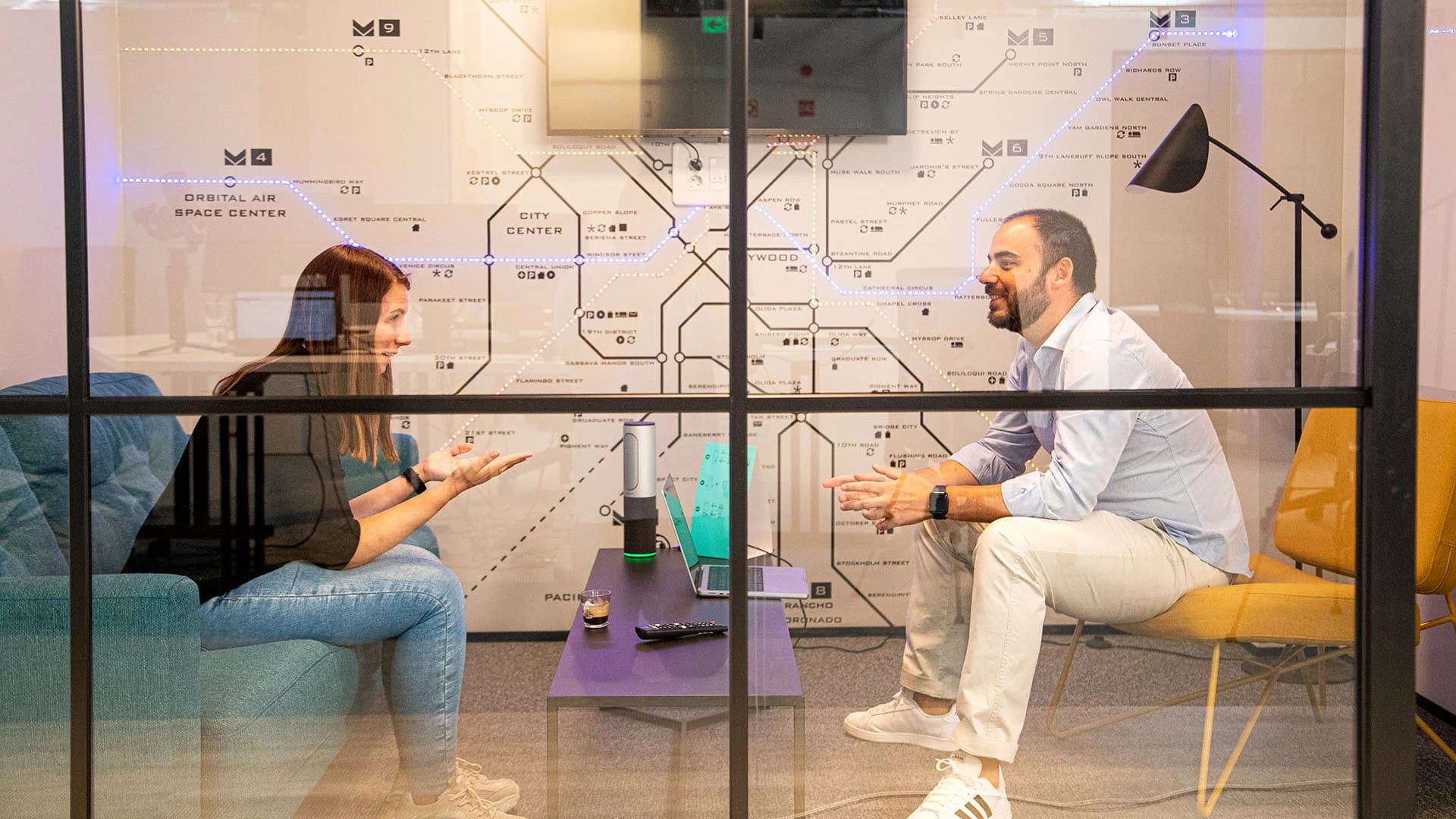 In a room, two people are seated with a map on the wall, engaged in a conversation about secure cybersecurity infrastructure