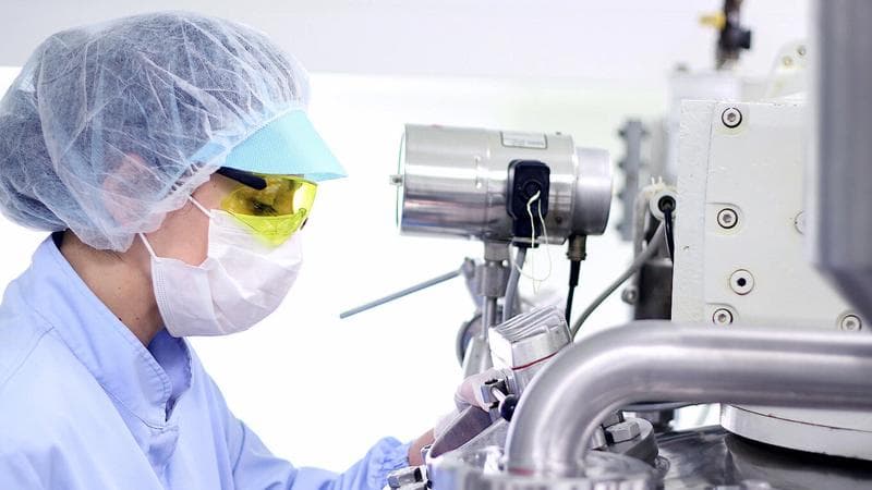 Frau mit weißem Laborkittel und Brille bedient eine Maschine.