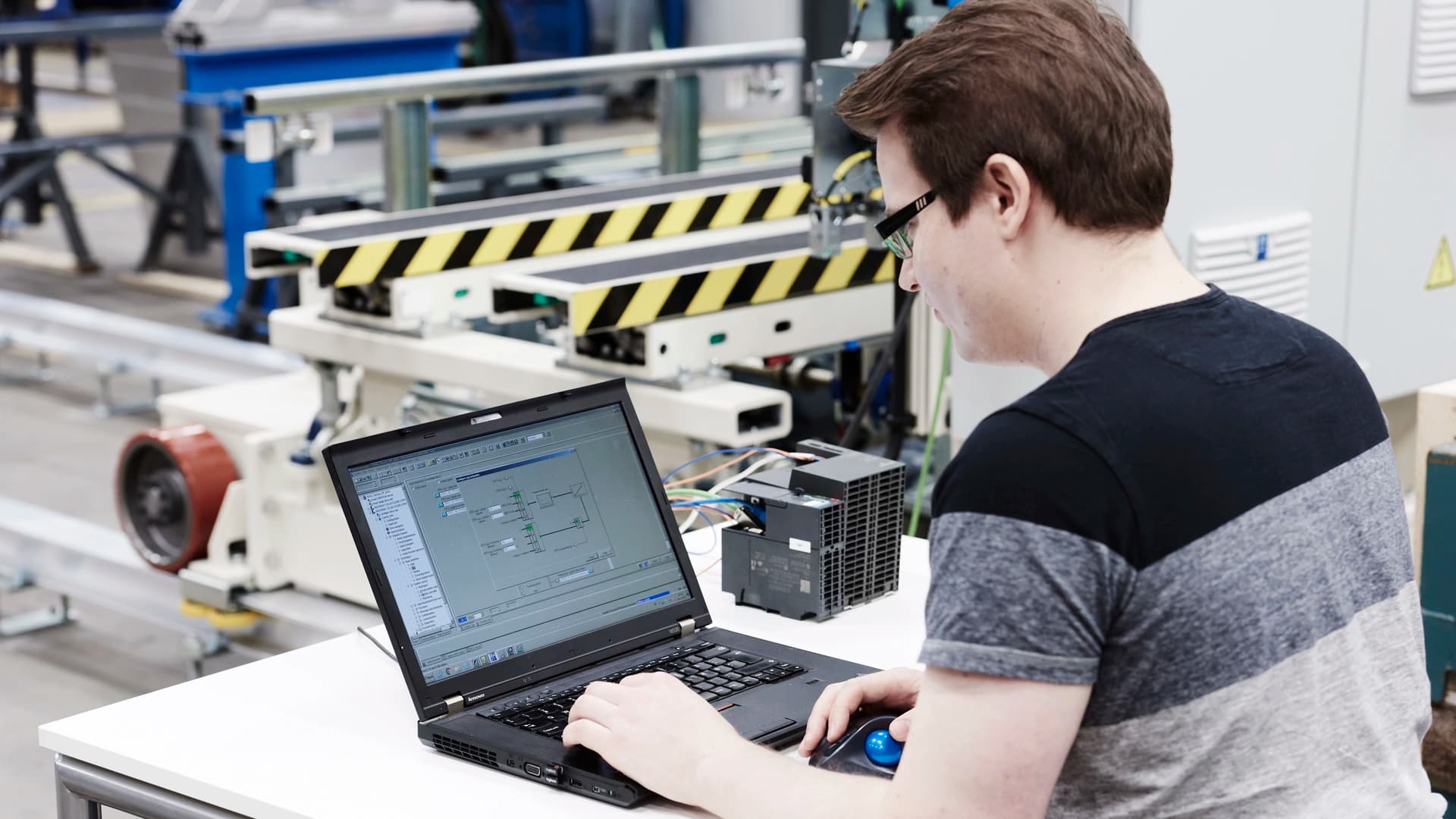 A man focused on a laptop while seated at a workstation in a factory environment.