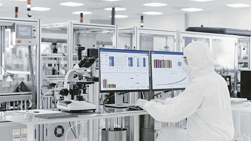 Laboratory worker, wearing a hazmat suit and looking at data in two screens, in a laboratory setting.