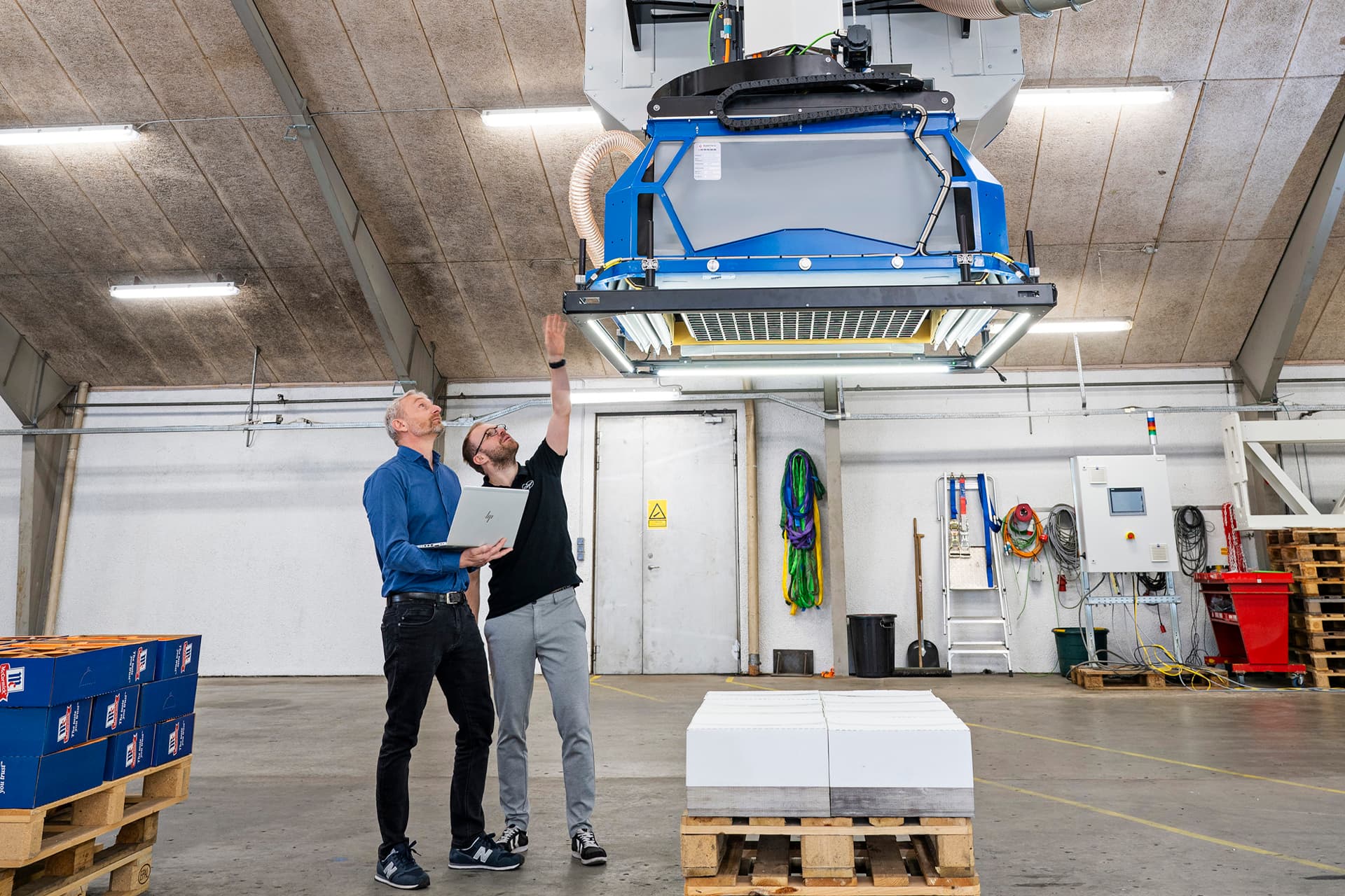 Zwei Ingenieure inspizieren ein automatisiertes Überkopf-Handlingsystem in einem Lager und diskutieren über eine schnellere und nachhaltigere Logistiklösung.