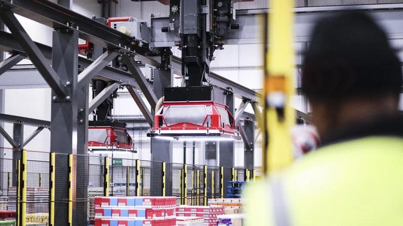 A worker stands in front of a large red and white truck, showcasing modern warehouse automation and layer picker technology.