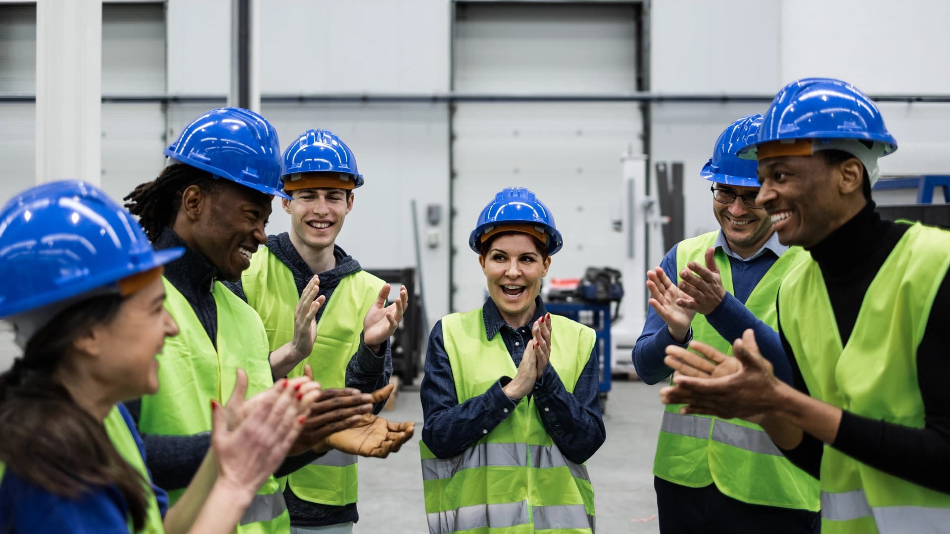 A group of workers wearing hard hats and vests enthusiastically clapping together in a construction setting.