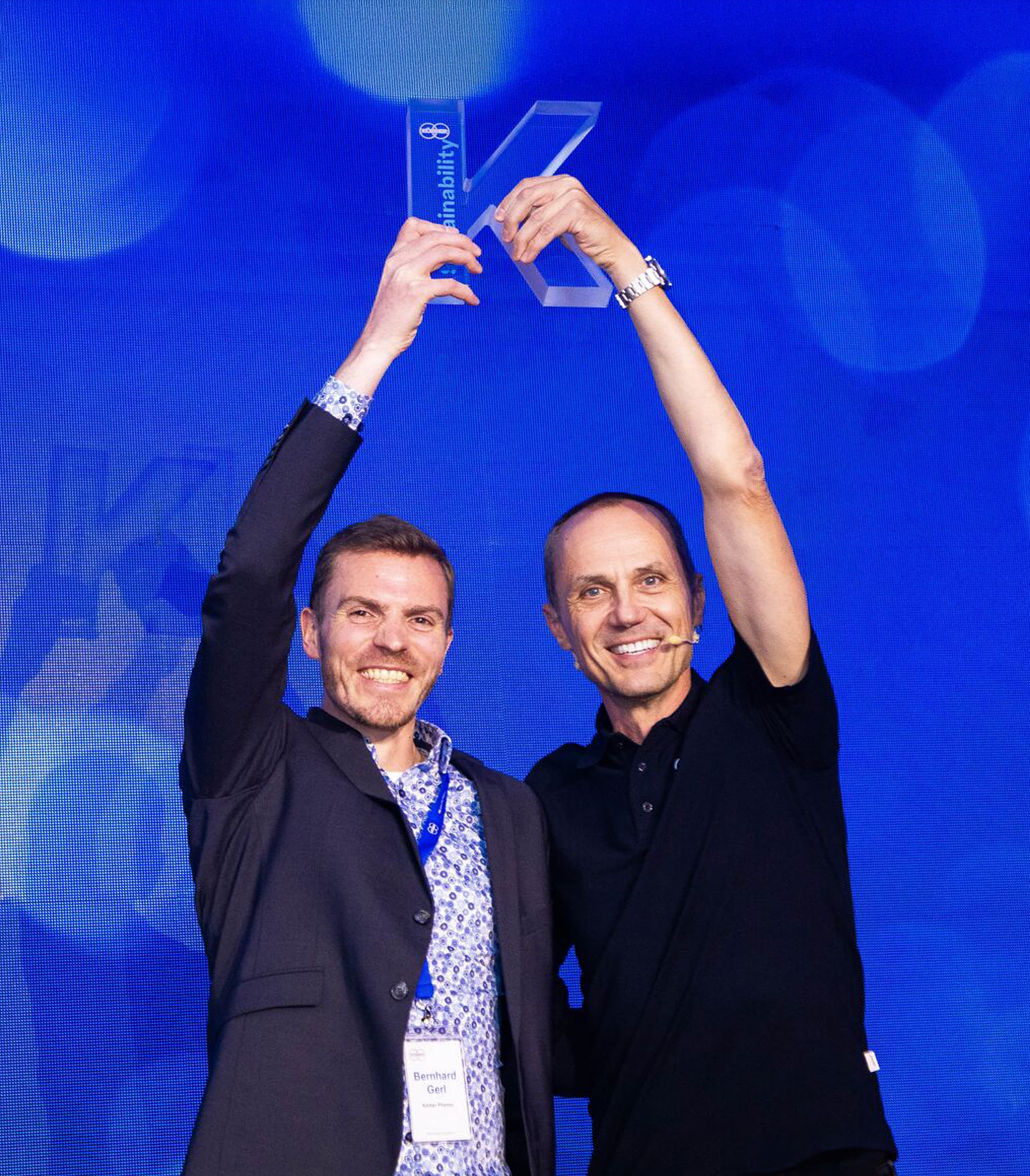Two male Körber employees holding a glass aware in the shape of a K about their heads