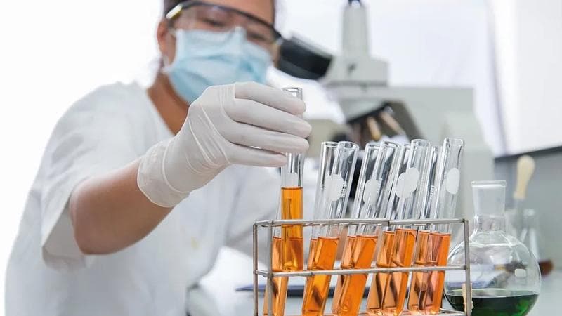 Person mit Handschuhen, Maske und Schutzbrille, die ein Reagenzglas mit einer orangefarbenen Flüssigkeit darin aufnimmt.