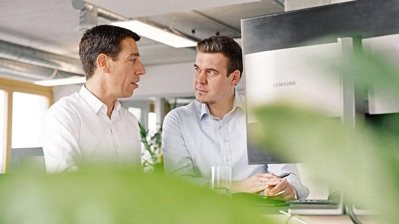 Two men sitting in an office setting, talking to each other.
