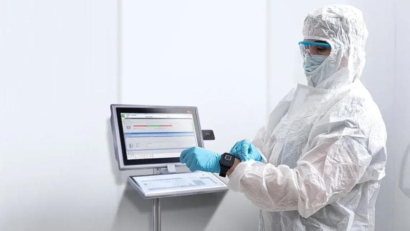 Eine Person in Labor-Schutzkleidung, wie einem Schutzanzug, einer Schutzbrille und Handschuhen, sitzt neben einem Computer.
