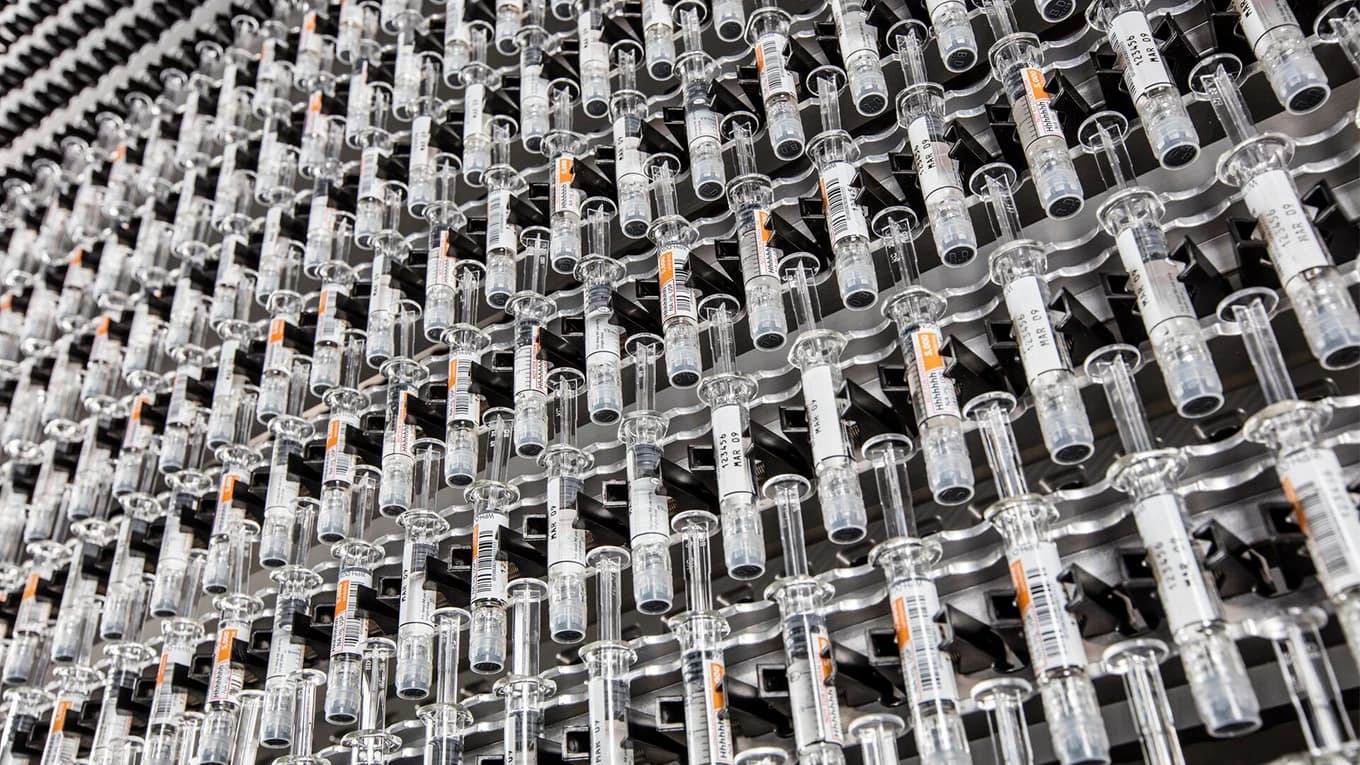 Glass vials with needles arranged in a row, representing mass production in the pharmaceutical industry.