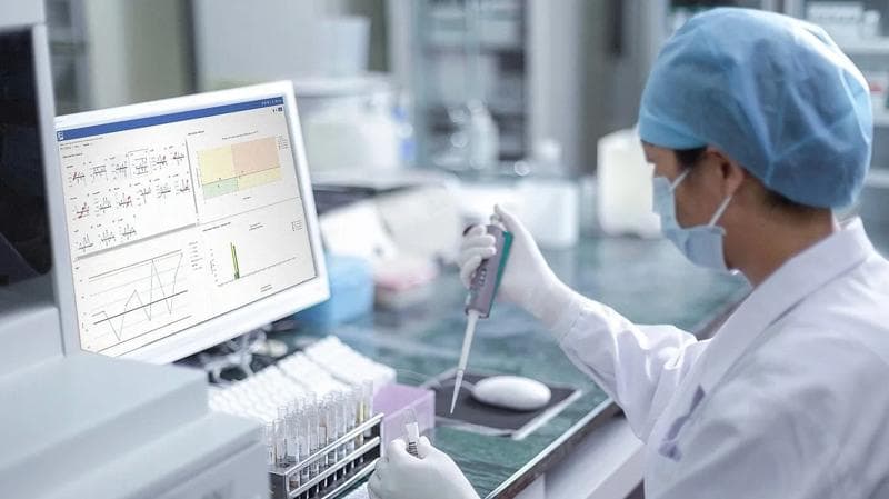 Person wearing medical protective gear,looking at a computer screen, analyzing substances in test tubes.