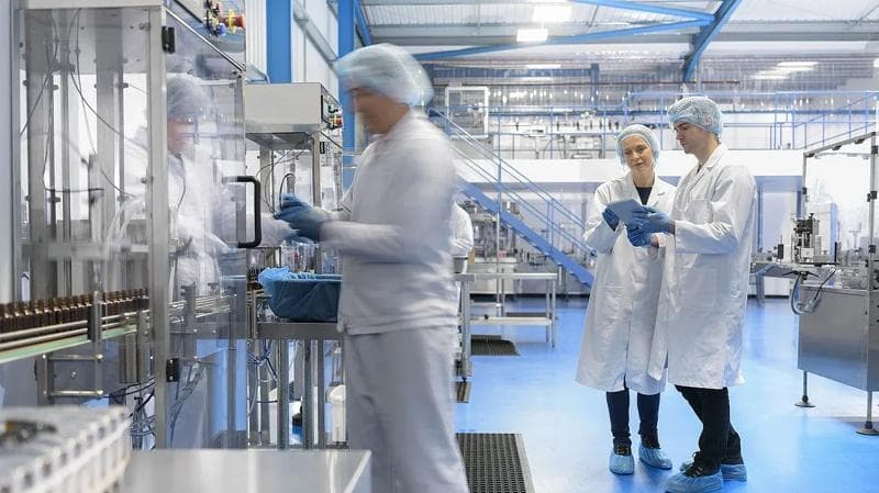 Laboratory workers, wearing medical protective gear, in an industrial setting, surrounded by pharma machines.