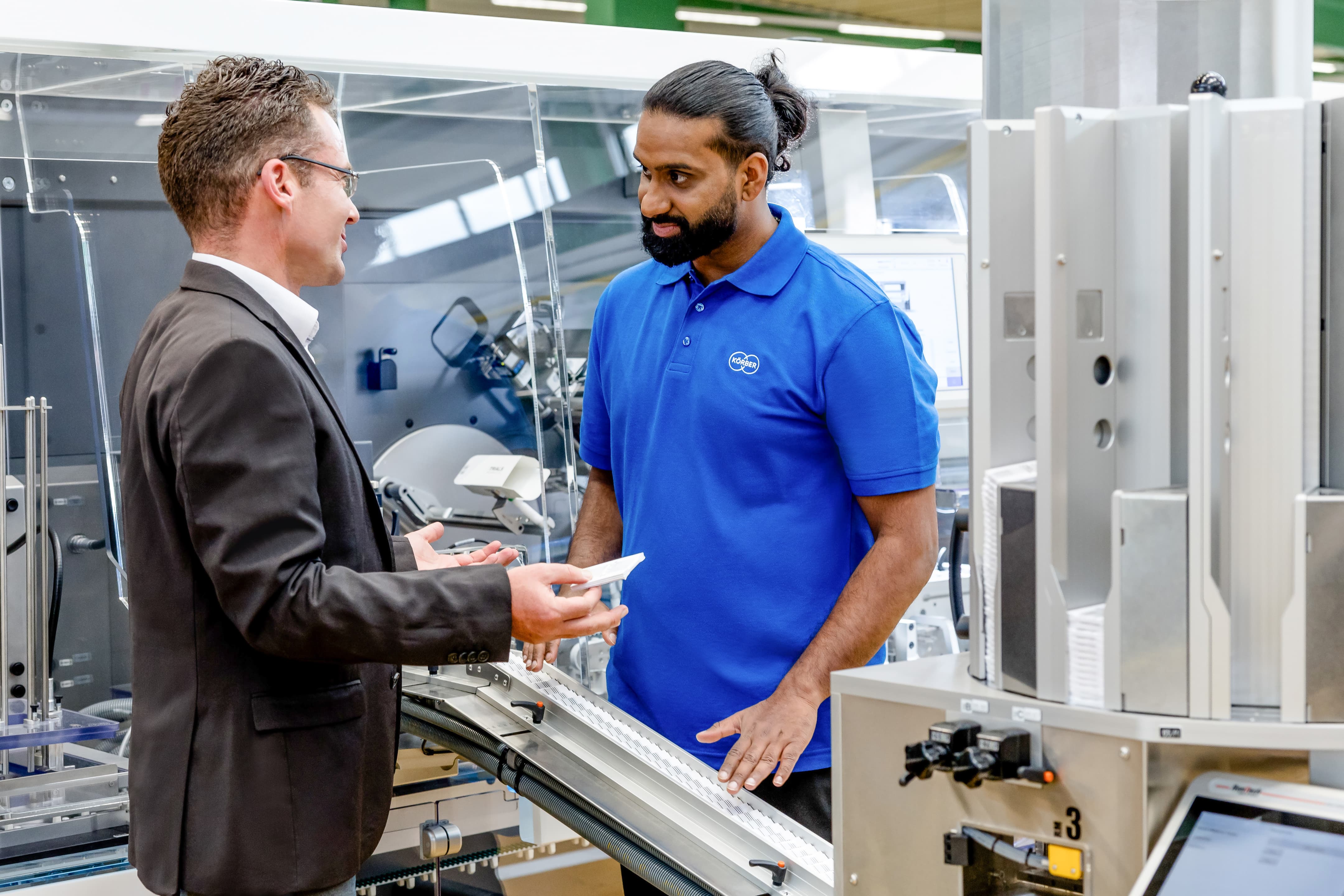 Two men in a Körber factory. One of the holds a pharmaceutical leaflet.