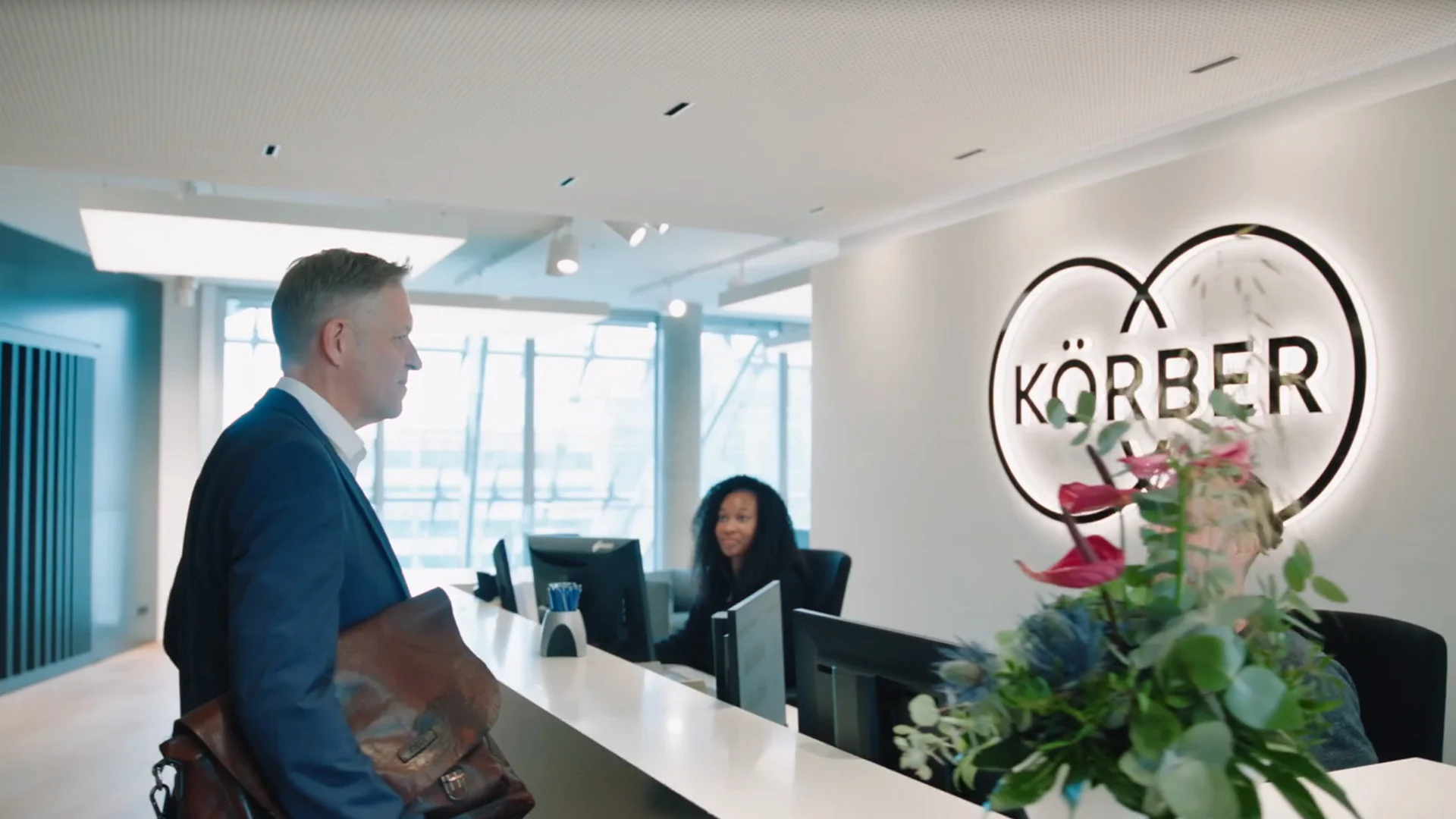 A visitor arrives at the Körber reception desk, with the illuminated Körber logo visible behind the receptionist.