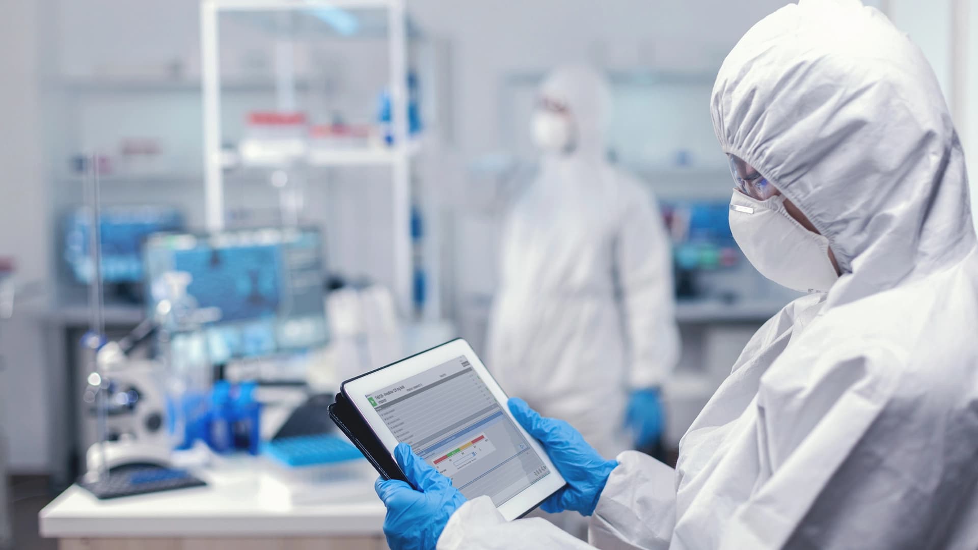 Person in einer Laborumgebung mit Laborkittel und Maske, die ein Tablet benutzt.