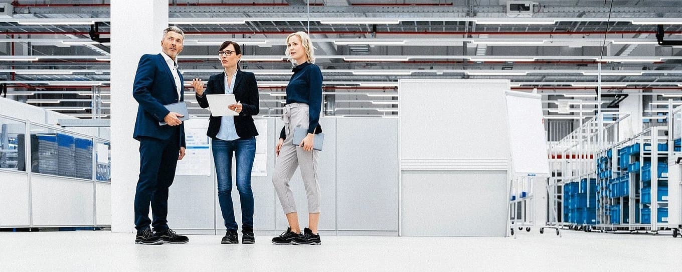 Drei Geschäftsleute stehen zusammen in einem Industriegebiet, umgeben von Maschinen und Lagerhallen.