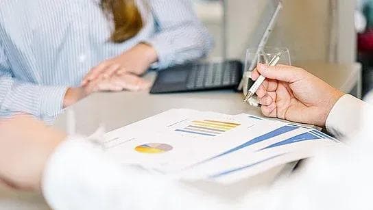 Zwei Personen an einem Tisch, die an Unterlagen und Computern arbeiten.