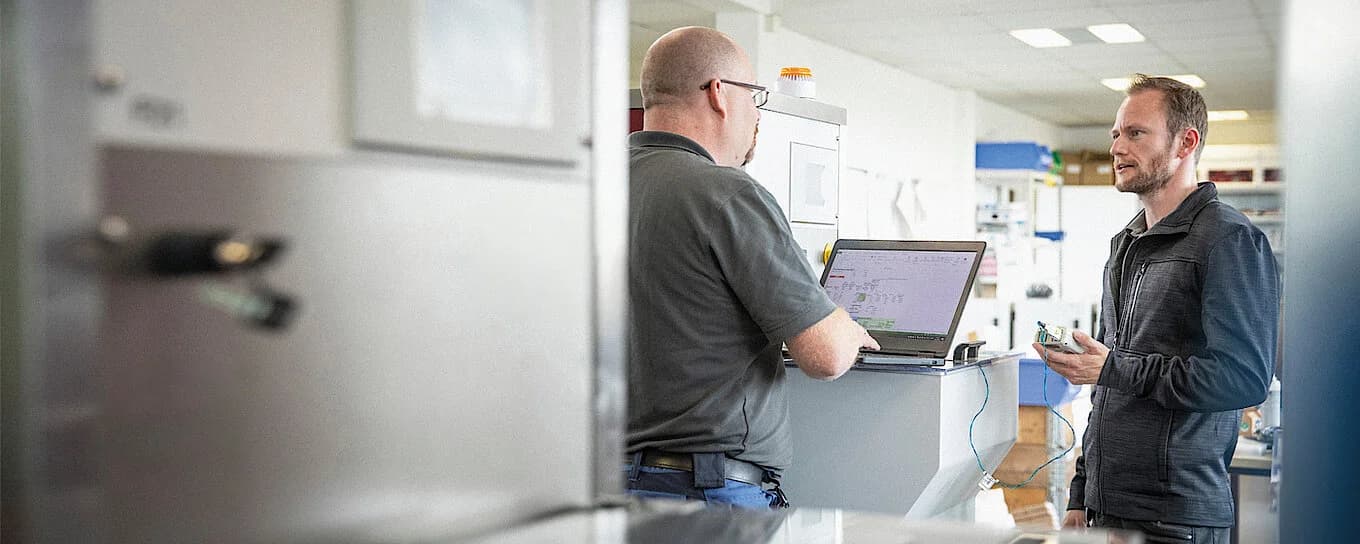 Two man talking while working - one on a computer and the other one with a small machine in his hands.