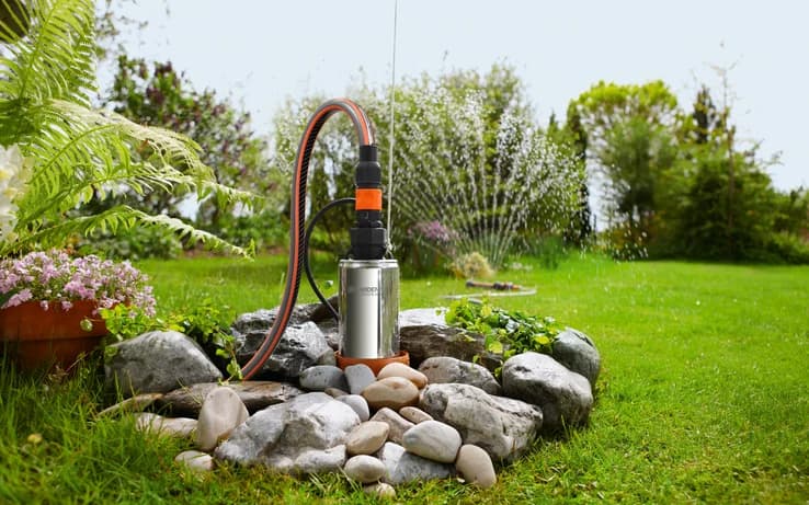 A water pump surrounded by rocks and various plants in a lush garden setting.