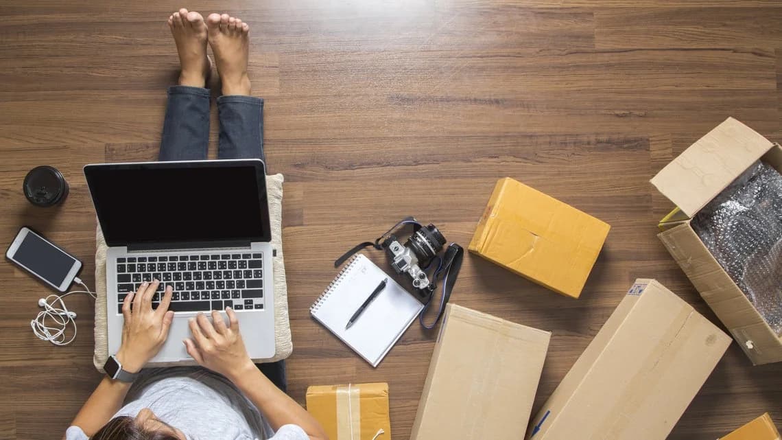 A person sits on a wooden floor with a laptop and several boxes around them.
