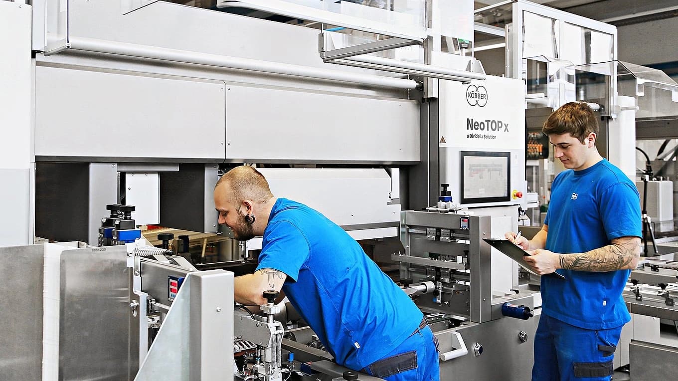 Two men operating a machine in a factory setting, focused on their tasks amidst industrial equipment.