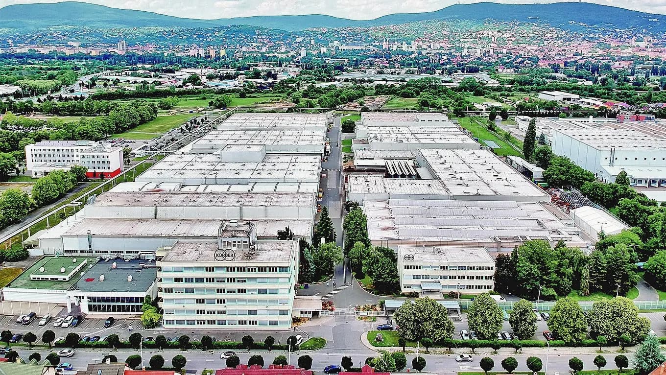Luftaufnahme der hochmodernen Produktionsstätte von Körber auf dem Campus des Unternehmens in Pécs, Ungarn, umgeben von Grünflächen und Industriegebäuden.