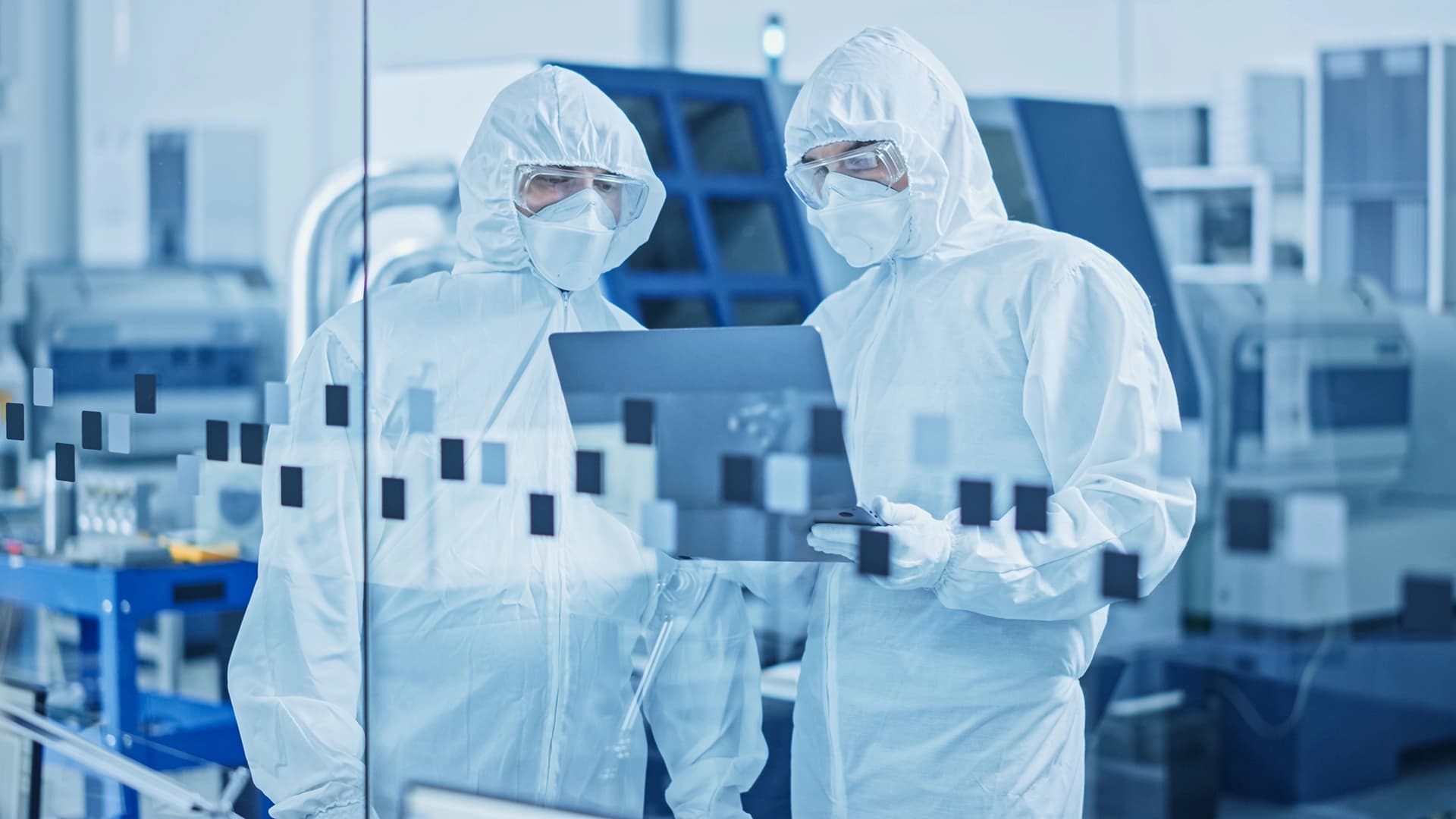 Zwei Personen in weißen Schutzanzügen analysieren Daten auf einem Laptop in einer pharmazeutischen Produktionsumgebung.