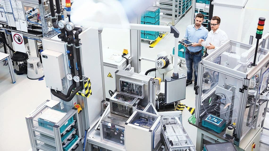 Two men are positioned in a factory, with various machines operating in the background.