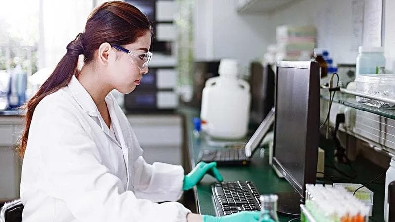Woman in a lab coat wearing goggles and looking at a microscope.