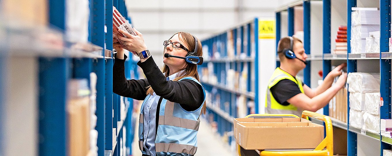 Männer und Frauen stellen in einem Warenlager Bestellungen zusammen, sie tragen Headsets, über die sie Arbeitsanweisungen bekommen.