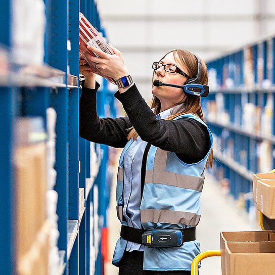 Männer und Frauen stellen in einem Warenlager Bestellungen zusammen, sie tragen Headsets, über die sie Arbeitsanweisungen bekommen.