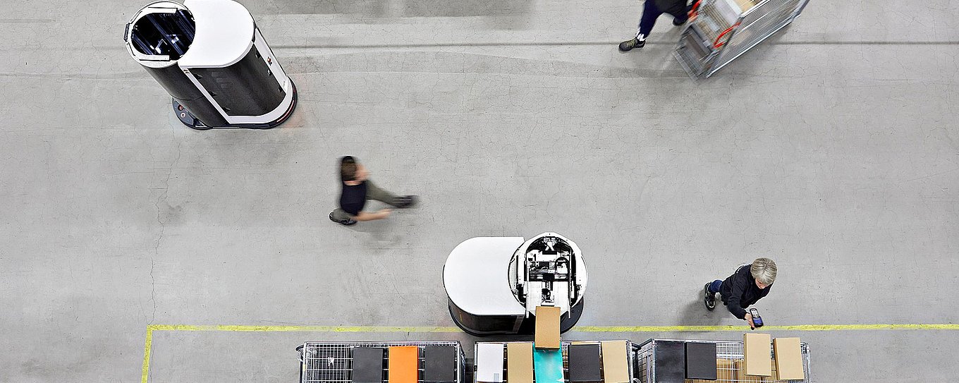 Aerial view of two Toru robots moving between workers in a high-bay warehouse.