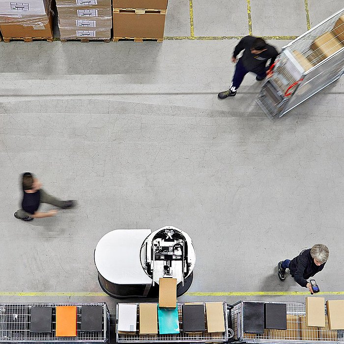 Aerial view of two Toru robots moving between workers in a high-bay warehouse.